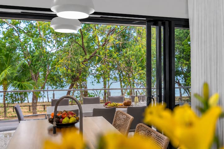 Dining table in the upper-level open-plan living area, with glass doors open to the balcony and the sea beyond the trees.