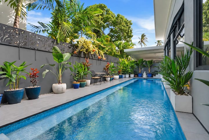 Long private saltwater pool beside a paved terrace lined with potted tropical plants.