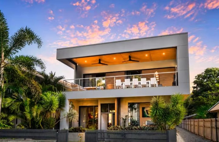 The two-storey house at dusk, with its lit upper balcony, palms and front lawn.