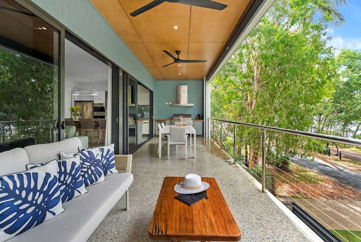 Covered balcony with a daybed and timber coffee table, overlooking the trees towards the sea.